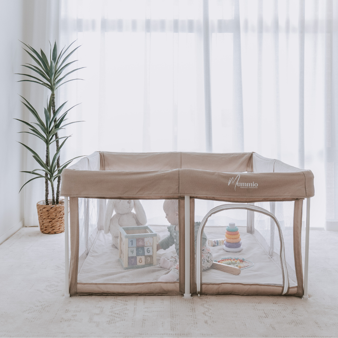 A beige Portable Safe Space baby playpen with clear mesh sides, set up in a bright nursery with sheer white curtains, a potted plant, and baby toys inside.