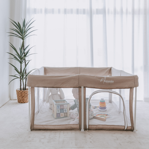 A beige Portable Safe Space baby playpen with clear mesh sides, set up in a bright nursery with sheer white curtains, a potted plant, and baby toys inside.