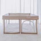 A Mummio Portable Safe Space dune baby playpen with a white mesh canopy and a small attached arch play door, set up on a light-colored floor in front of white sheer curtains. The brand name 'Mummio' is printed on the side.