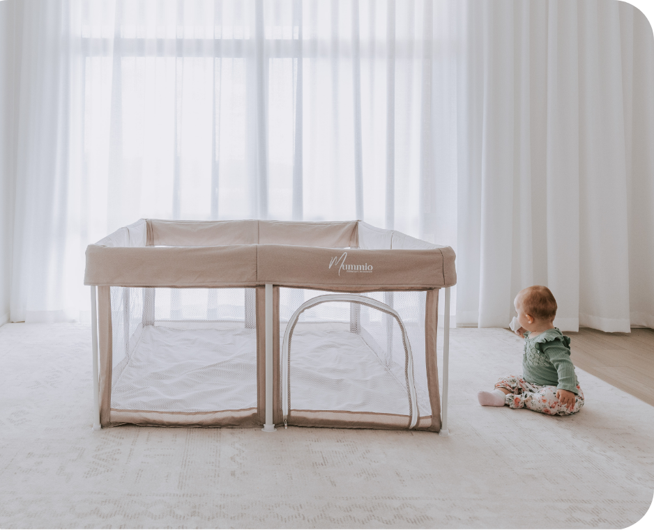 A Portable Safe Space baby playpen with a dune canopy and mesh sides, set up on a light-colored rug in a bright room with sheer white curtains. A baby in a green outfit is sitting on the floor to the right of the playpen.