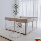 Beige dune colour portable pop up play pen with a white frame, set up on a light coloured mat. In a bright lit room with white sheer curtains and a green potted plant. 