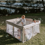 Mummio portable pop up playpen for outdoor use. Dune in colour with a white frame. Four kids are happily playing inside. There are bubbles in the air. It is on grass with a lake behind 