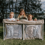 Portable baby play pen in Dune with four children inside smiling. The brand “Mummio” is printed on the front of the play door. Outdoor playpen with a lake behind. 