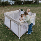 Mummio pop up portable playpen outdoors with four children playing. All different ages - 11 months, 2 years, 3 years and 4 years old children. They are on grass with the lake in the background 