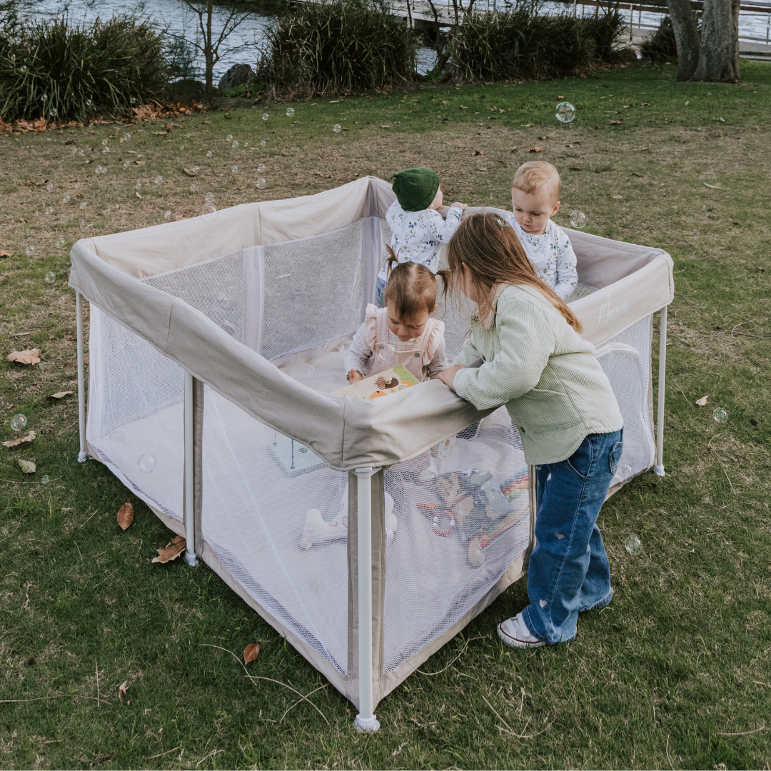 Mummio pop up portable playpen outdoors with four children playing. All different ages - 11 months, 2 years, 3 years and 4 years old children. They are on grass with the lake in the background 