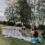 Portable pop up baby and toddler play pen. Mums and children outdoors having a picnic on the grass with a lake behind. Therr is a toddler balance bike and a pram 