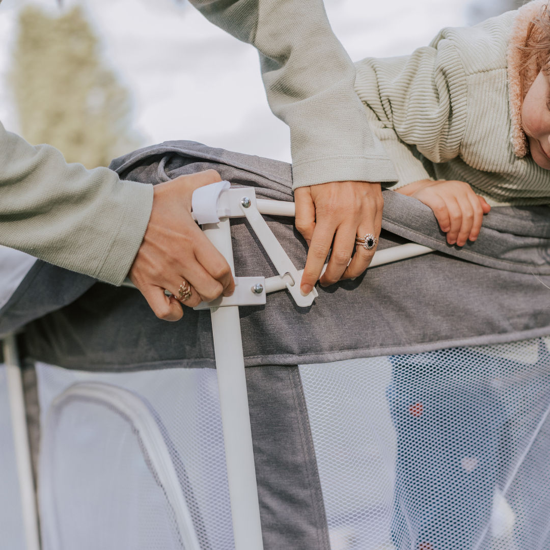 Portable Safe Space play pen one latch locking. Safety lock for pop up play pen. Woman setting it up and child watching. Stone grey play pen with a white frame