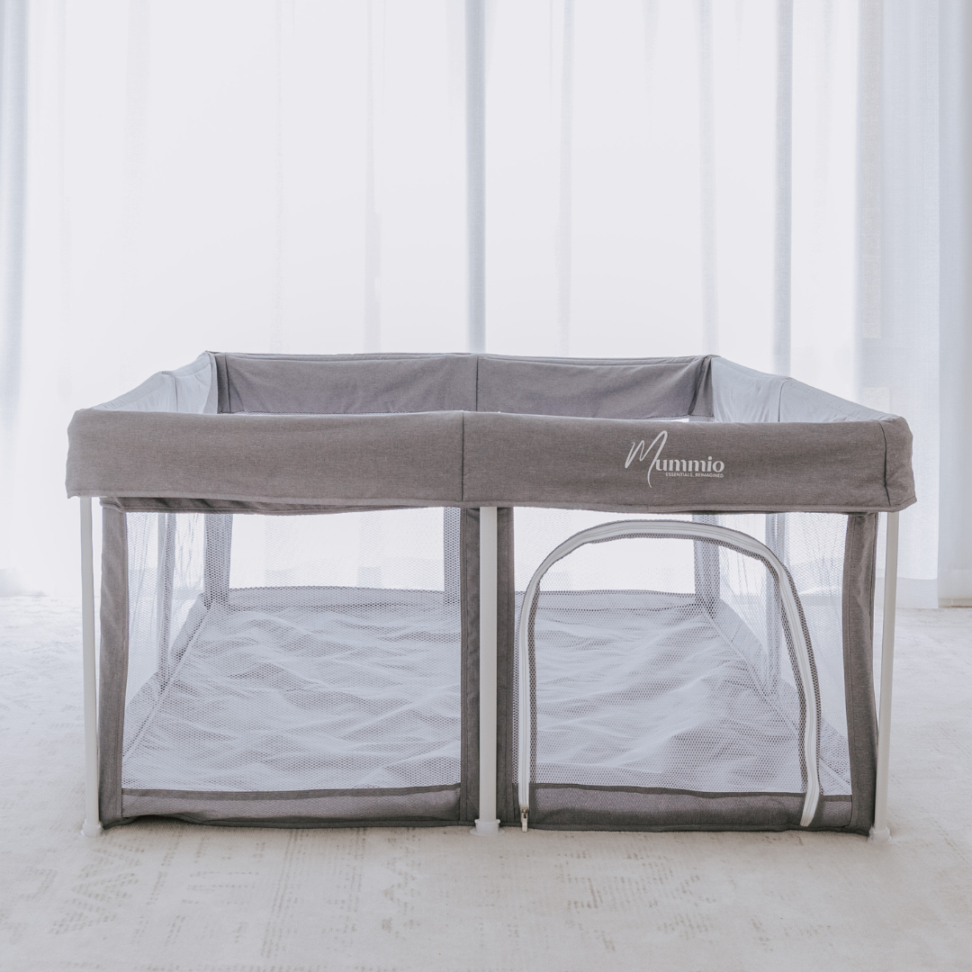 Grey stone pop up portable play pen with a white frame and the mummio logo on the front. Set up on a light mat with a light sheer curtain. Easy toddler play door.