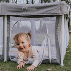 Stone grey pop up portable play pen with a white frame. Young girl crawling out of the play door smiling. The "mummio" brand logo is on the front of the play pen.