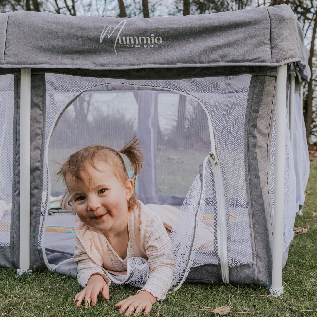 Stone grey pop up portable play pen with a white frame. Young girl crawling out of the play door smiling. The "mummio" brand logo is on the front of the play pen.