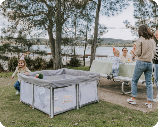 A gray and white outdoor baby playpen is set up on a grassy lawn next to a picnic table with people and children in a park by the water. The Portable Safe Space party